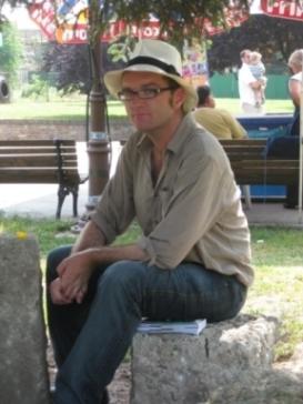 James Kennaway wearing a hat and siting on a stone in a park