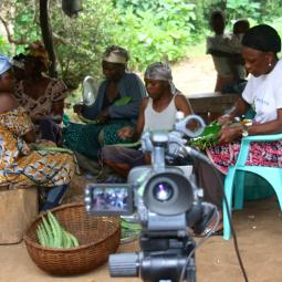 A video camera in front of women sat on chairs and stools